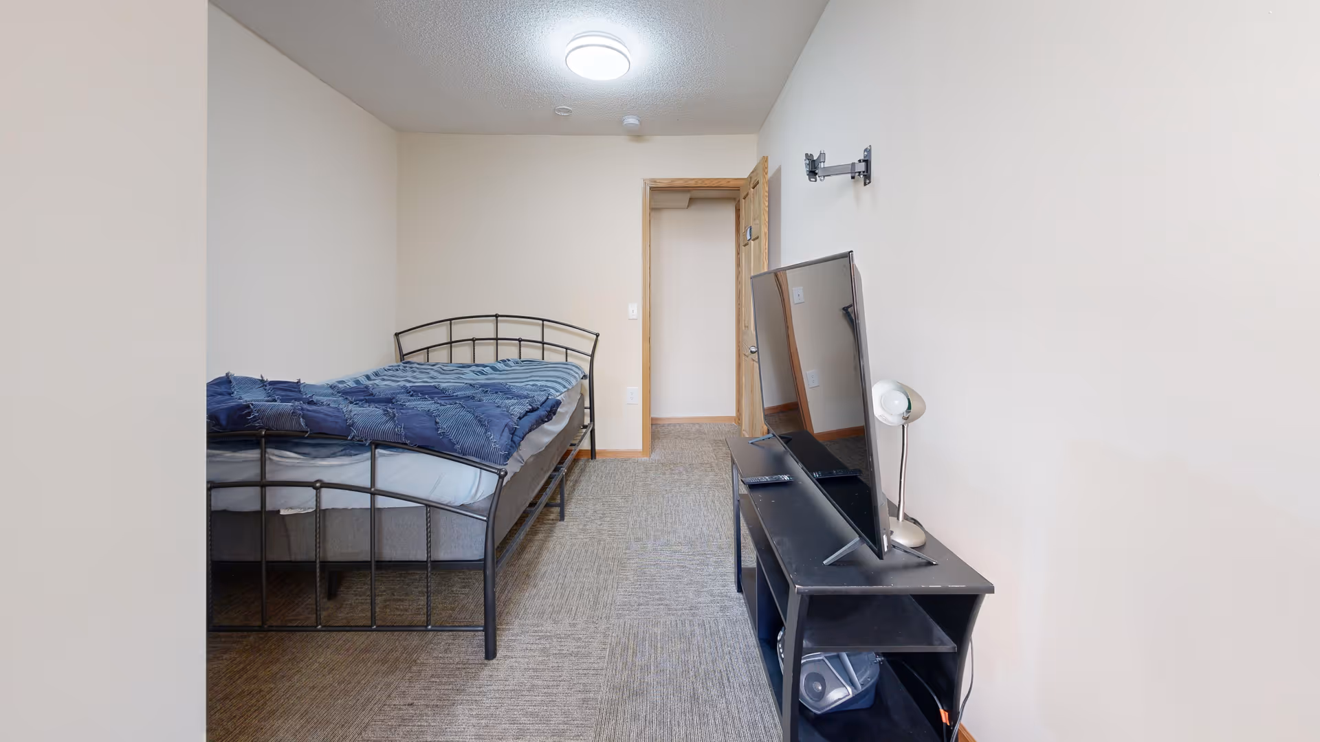 A simple bedroom with a metal frame bed covered with a blue blanket, a black TV stand holding a flat-screen TV and a small lamp, beige walls, and a carpeted floor. There is an open wooden door at the far end of the room.