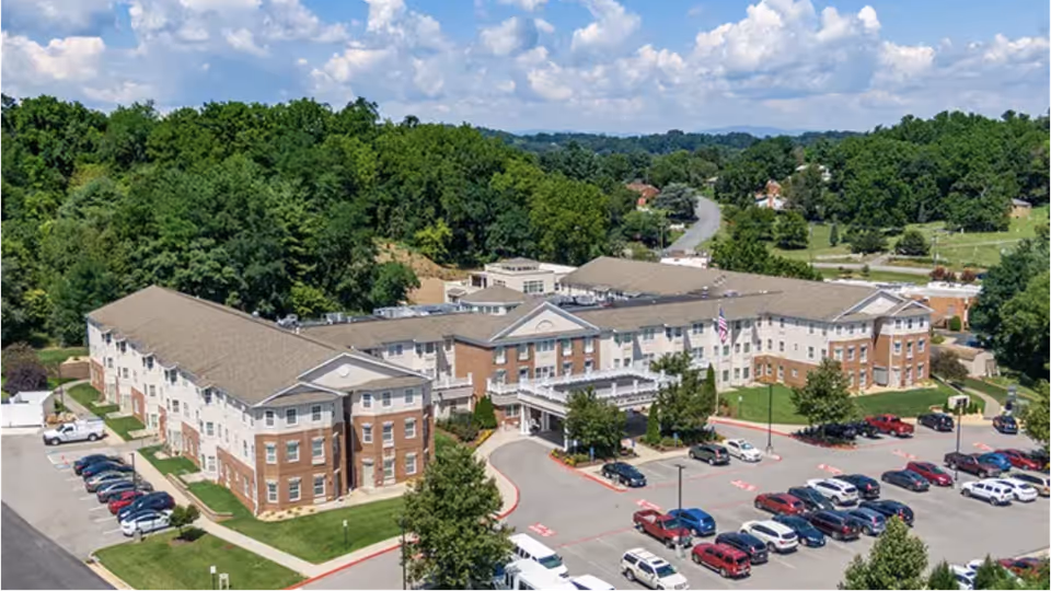 Aerial view of The Legacy at North Augusta senior living facility showing a large, multi-wing building surrounded by trees and greenery, with a parking lot filled with cars in front under a partly cloudy sky.