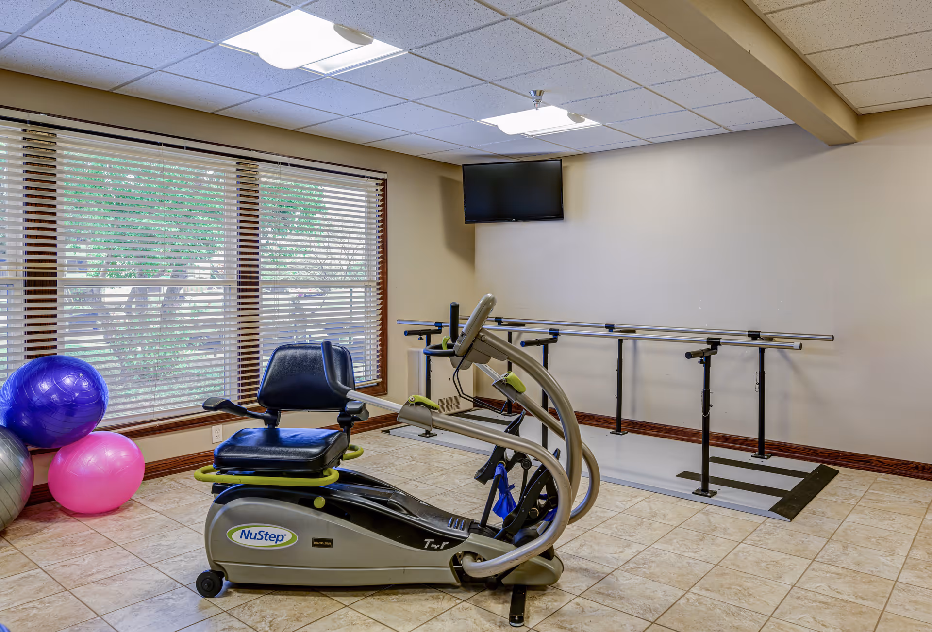 A small exercise room with a NuStep recumbent cross trainer, three exercise balls in purple, pink, and silver, a wall-mounted TV, and parallel bars for walking support. The room has beige walls, tiled floor, and a large window with blinds showing greenery outside.