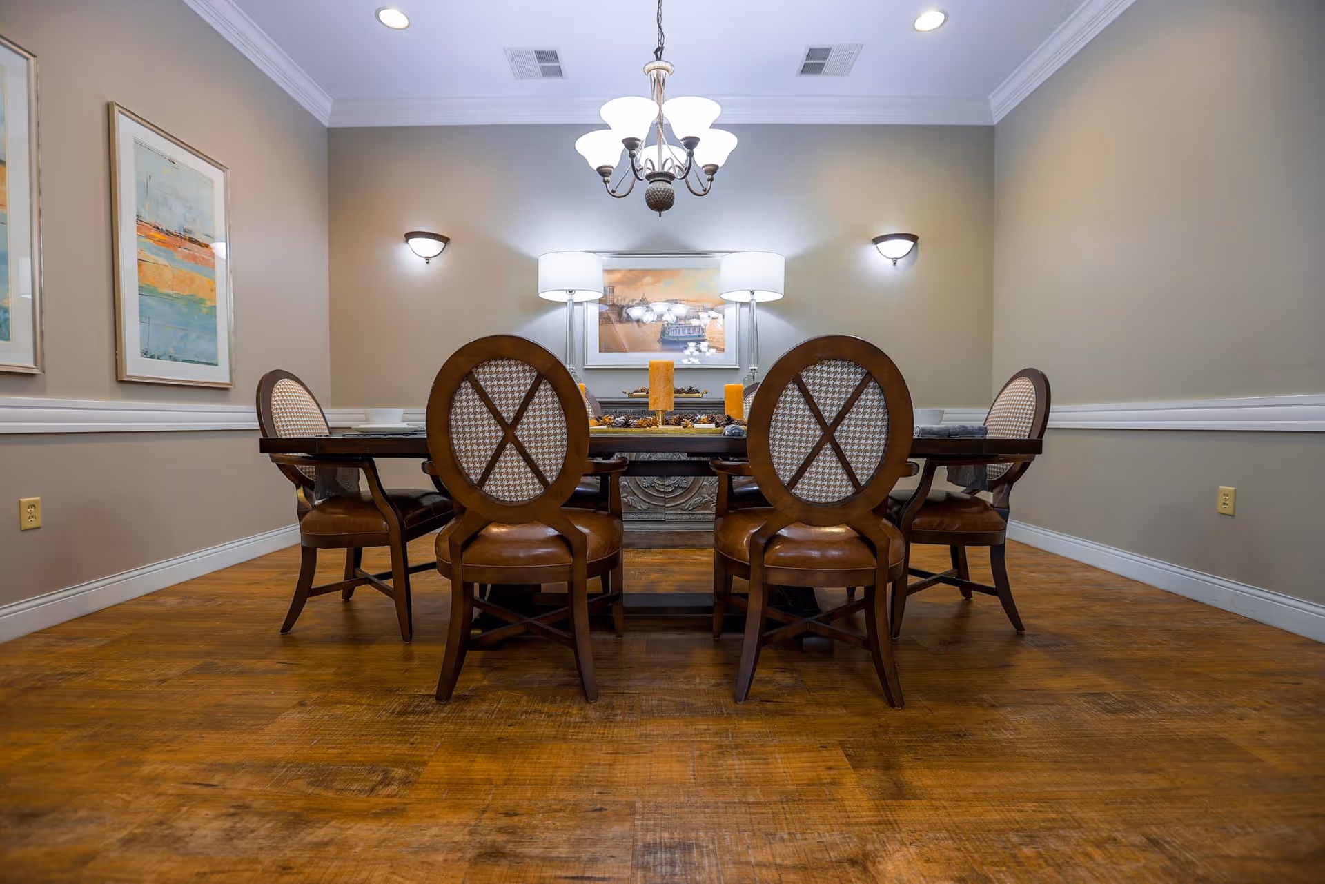 A dining room with a wooden table surrounded by six cushioned chairs with patterned backs. The room has wooden flooring, beige walls with white trim, two framed abstract paintings on the left wall, a chandelier hanging from the ceiling, two wall sconces, and two table lamps on a sideboard against the far wall.