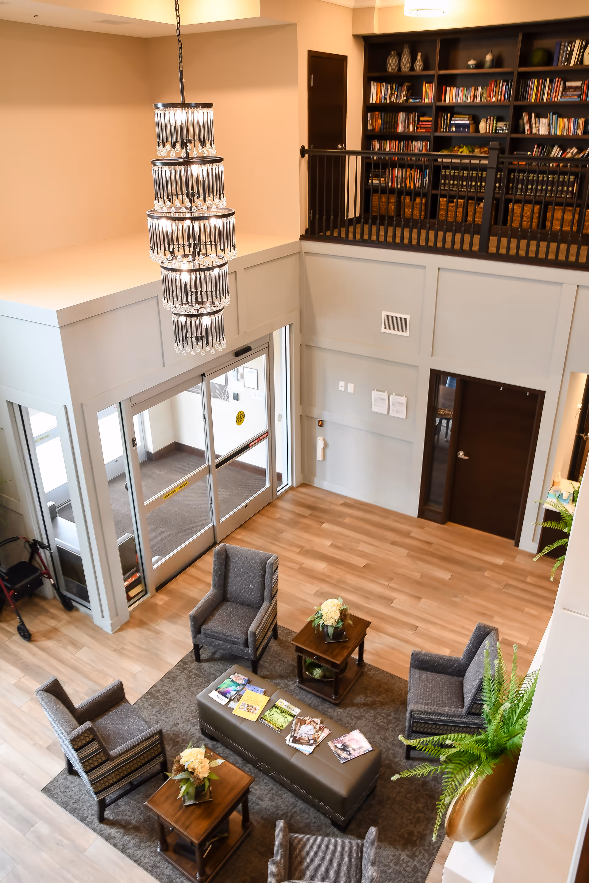 View from above of a senior living facility lobby with four gray armchairs arranged around a rectangular ottoman with magazines on it, two wooden side tables with flower arrangements, a large chandelier hanging from the ceiling, a bookshelf on the upper level, and glass entrance doors.
