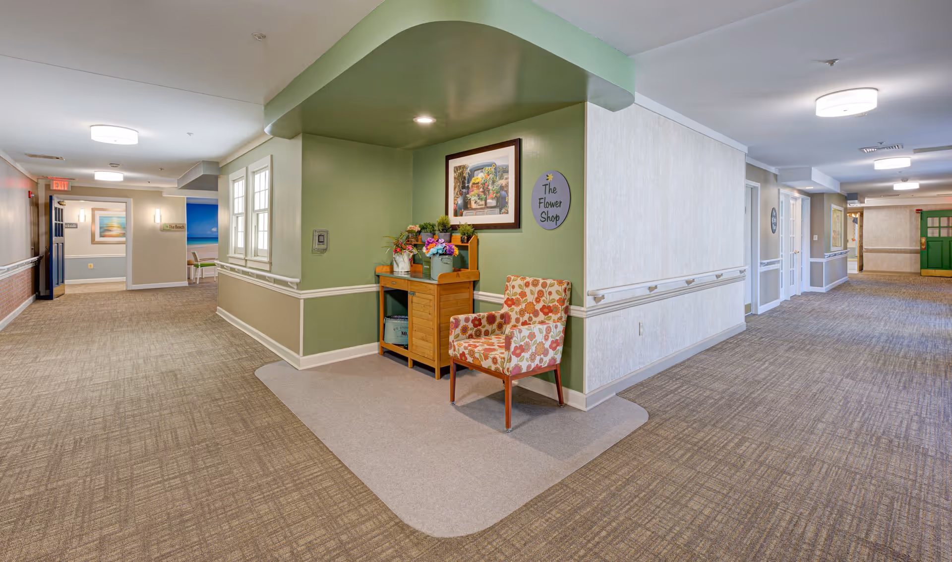 A well-lit hallway in a senior living facility with beige carpet and light green and white walls. There is a small nook with a wooden table holding flower arrangements and a floral-patterned armchair. A sign on the wall reads 'The Flower Shop.' The hallway extends in two directions with doors and framed artwork visible along the walls.