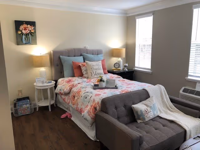 A cozy bedroom with a floral bedspread on a tufted headboard bed, decorative pillows including one that says 'Grateful Thankful Blessed', two bedside tables with lamps, a small gray loveseat with a pillow and a white throw blanket, wooden flooring, and two windows with blinds letting in natural light.