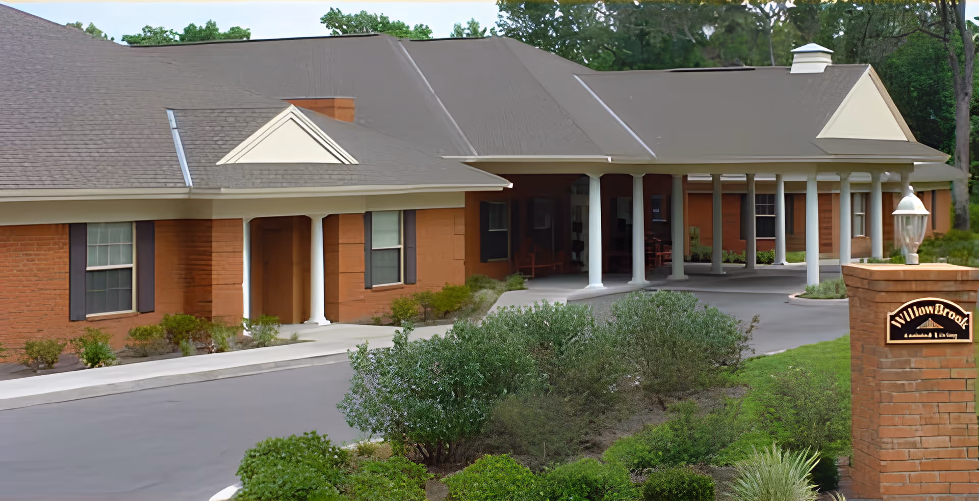 Front entrance of a brick assisted living building with a covered driveway, white columns, landscaping, and a brick sign reading "WillowBrook".