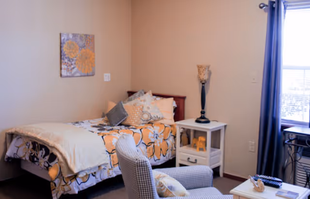 A cozy bedroom with a single bed covered in floral bedding and multiple pillows. There is a small white nightstand with a lamp and a decorative elephant figurine. A comfortable armchair with a pillow is positioned near a window with dark curtains, allowing natural light to enter the room. A floral wall art piece hangs above the bed.
