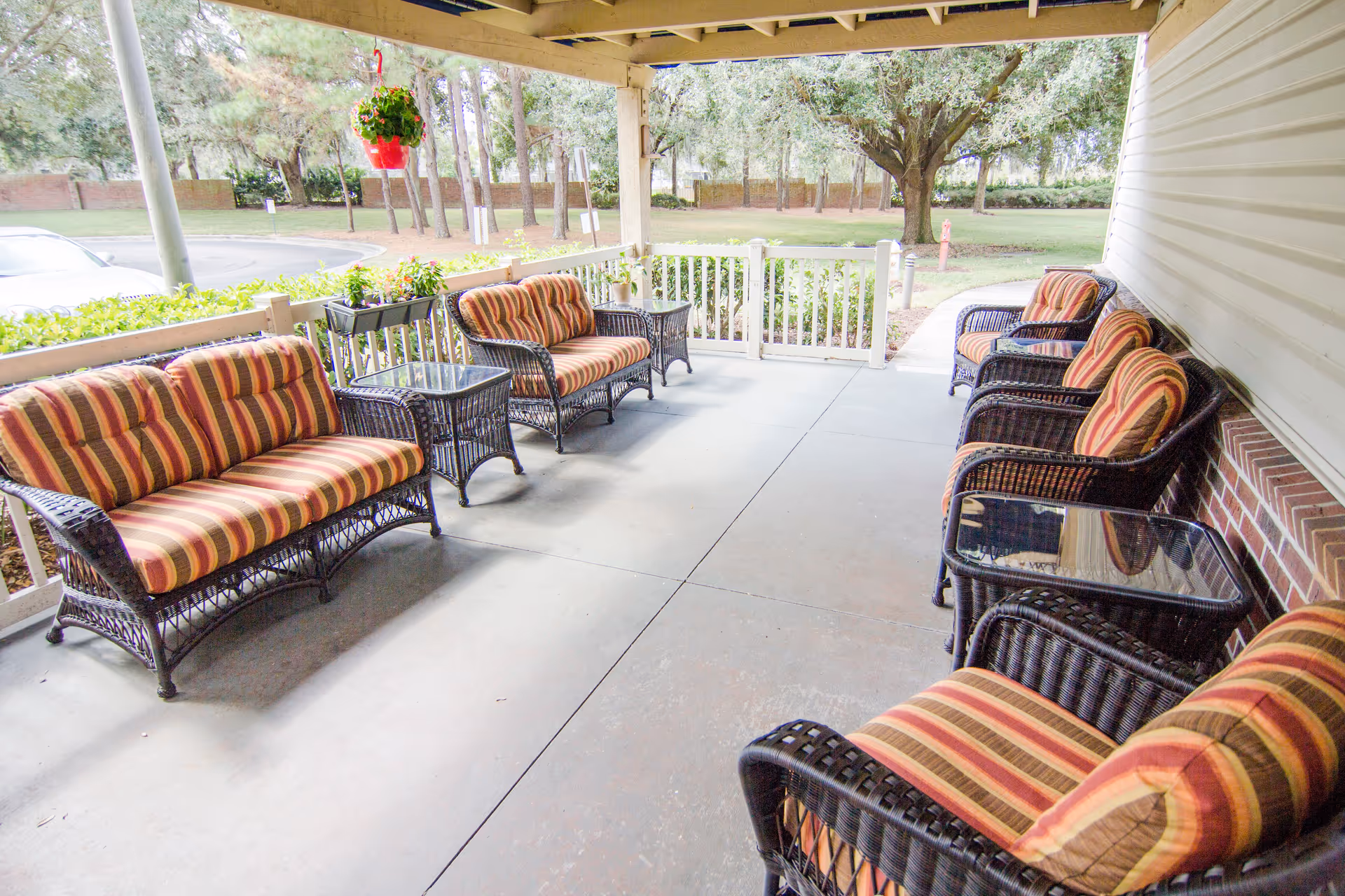 Covered outdoor patio area with multiple wicker chairs and loveseats featuring striped cushions in shades of red, orange, and yellow. There are glass-top side tables between the seating. The patio overlooks a grassy area with trees and a brick wall in the background. A hanging red flower pot with green plants is suspended from the ceiling.