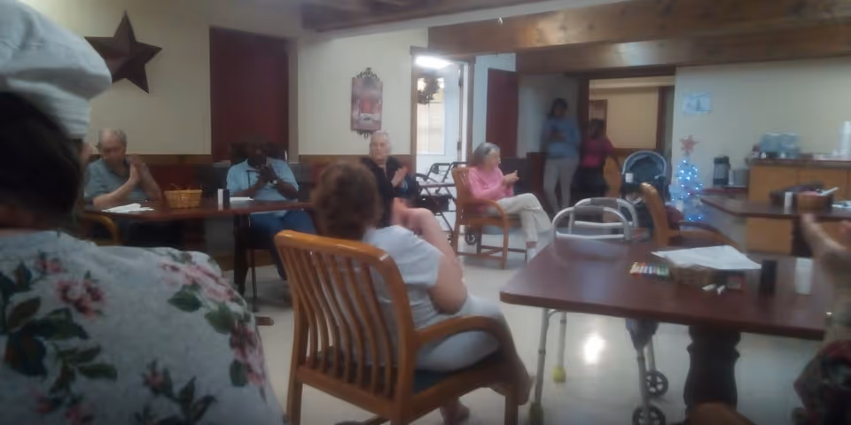 A group of seniors seated around tables in a communal activity/dining room with walkers and a small decorated tree.