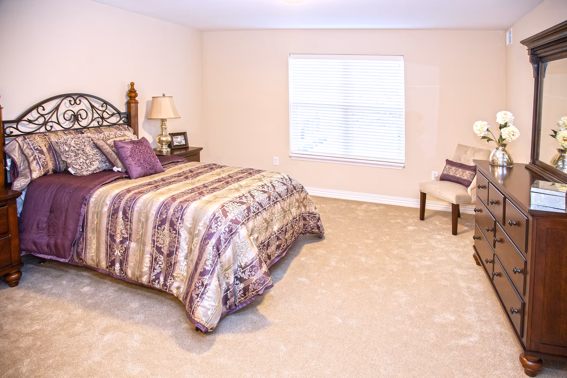 Bright bedroom with a decorative iron headboard, purple patterned bedding, a nightstand, dresser with mirror, and a chair by a window.