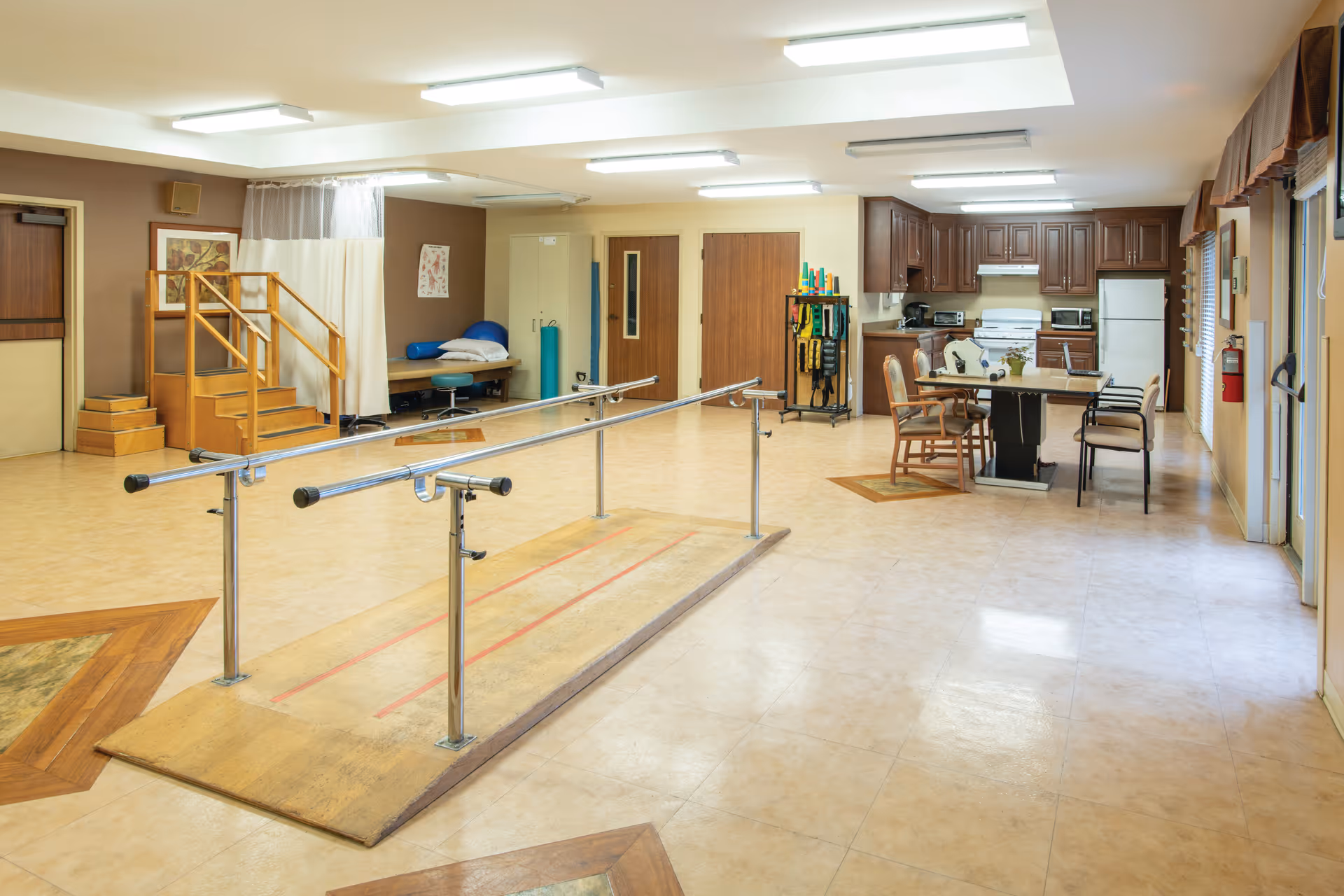 A spacious rehabilitation room with parallel bars for walking exercises in the center, a small set of wooden stairs with a handrail on the left, a treatment table with pillows and an oxygen tank against the back wall, and a kitchen area with wooden cabinets, a stove, microwave, refrigerator, and a table with chairs on the right side.