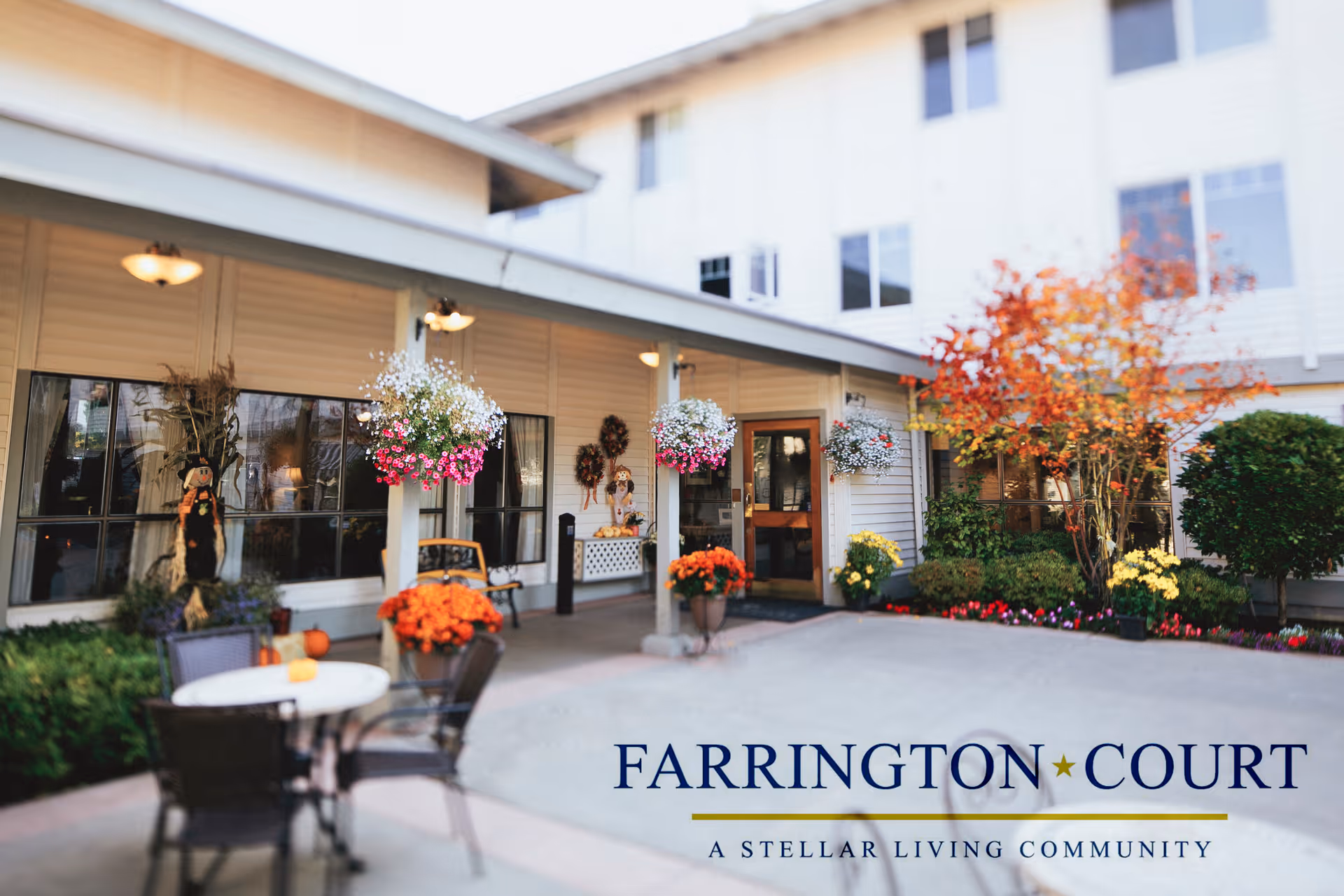 Outdoor patio area of a senior living community with a table and chairs, hanging flower baskets, potted plants, and autumn decorations near the entrance of a building.