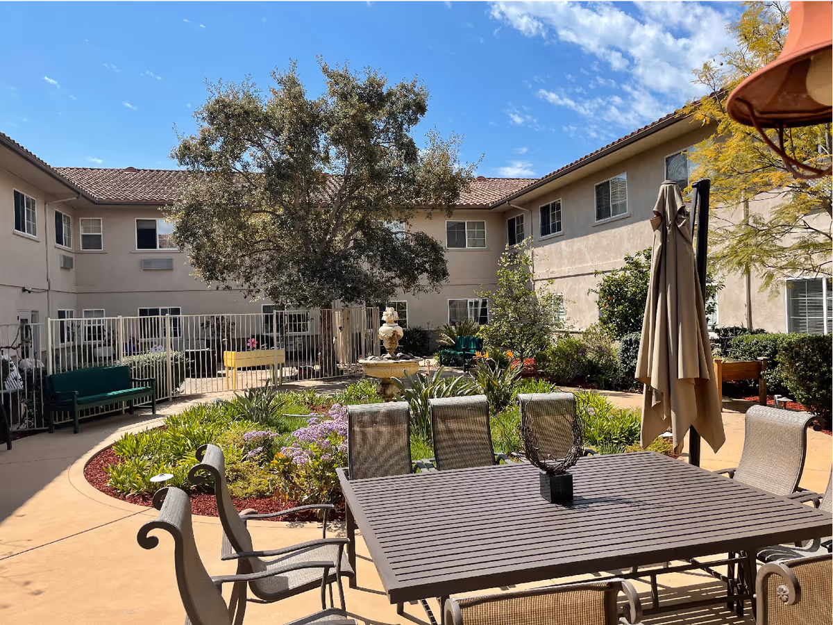 Sunny courtyard with a patio table and chairs, planted garden beds, a fountain and a surrounding two-story building.