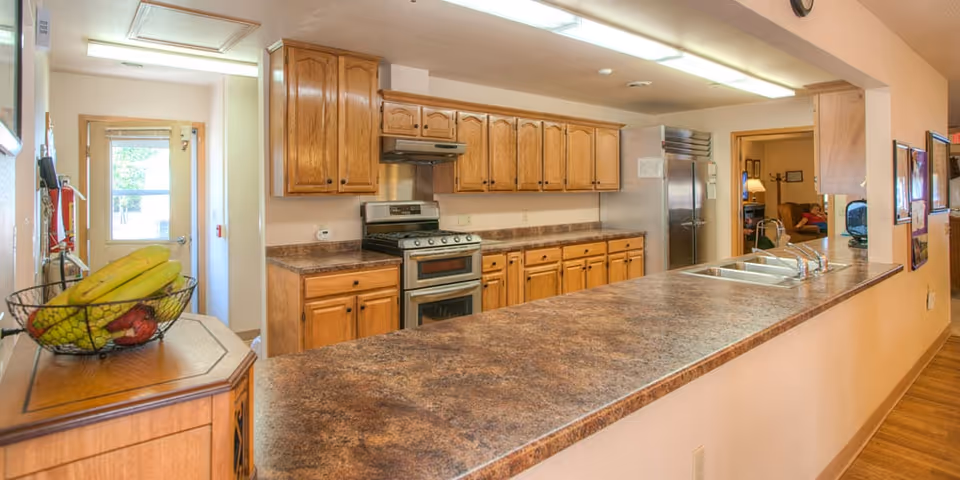 Long communal kitchen with wooden cabinets, a stove and refrigerator, double sink, and a long marbled countertop bar.