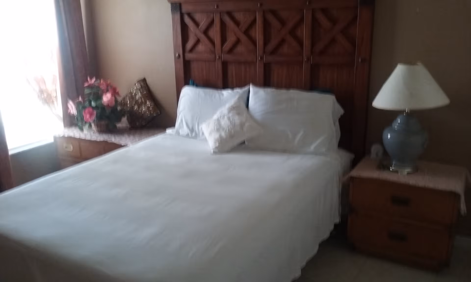 A tidy bedroom with a white-made bed, wooden headboard, nightstands, a lamp, and a floral arrangement by a window.
