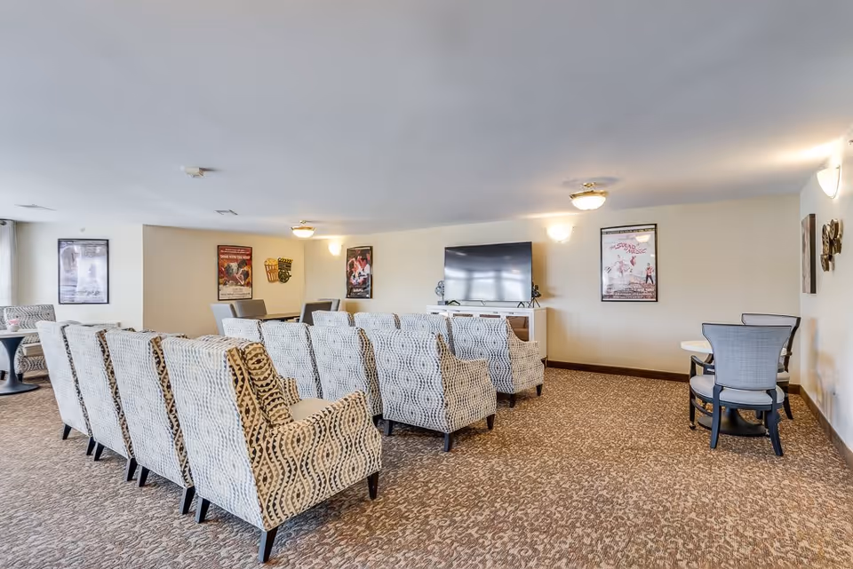 A spacious living room area with patterned armchairs arranged in rows facing a large flat-screen TV mounted on a white console. The room has carpeted flooring, light-colored walls adorned with framed posters, and additional seating with small tables and chairs along the sides.