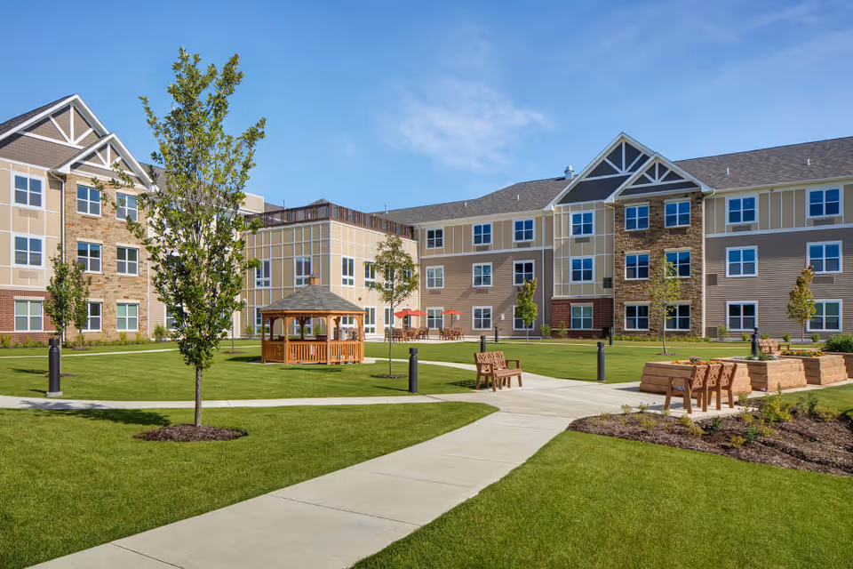 Outdoor courtyard area of a senior living facility with a well-maintained lawn, young trees, a wooden gazebo, benches, and raised garden beds, surrounded by a three-story building under a clear blue sky.