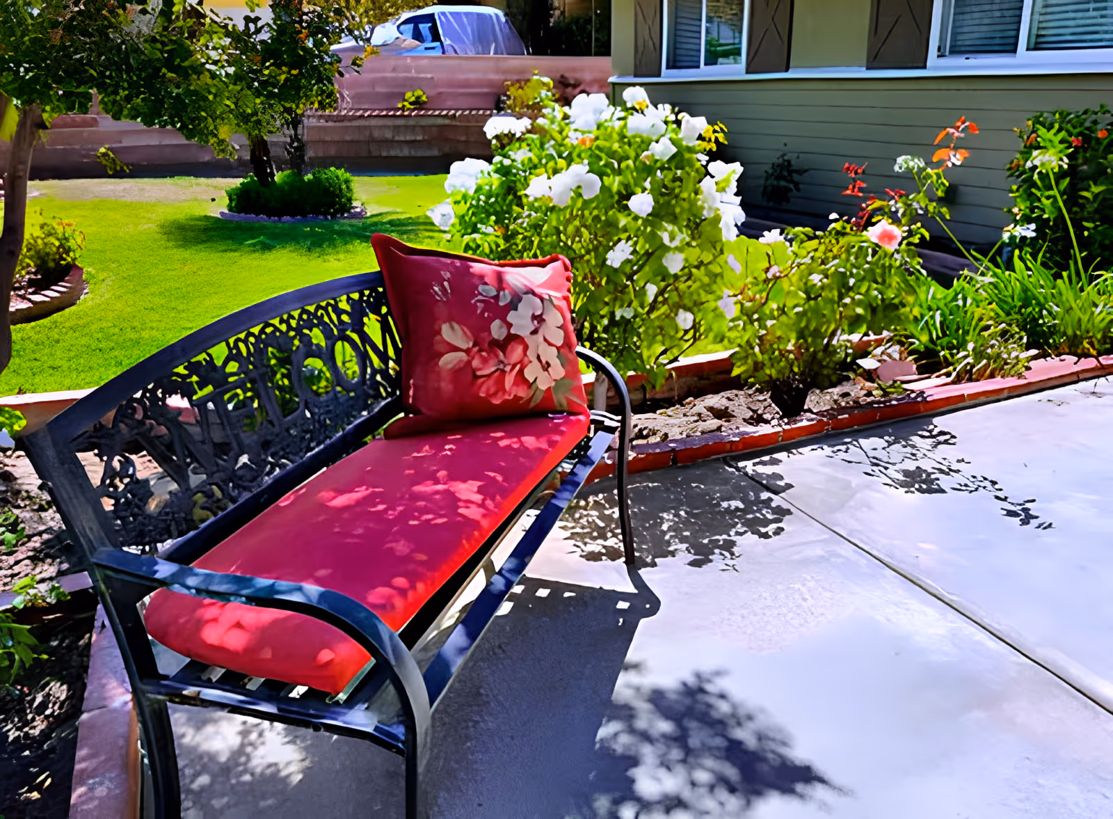 Outdoor garden area with a black metal bench featuring a red cushion and a floral pillow. The bench is placed on a concrete patio next to a flower bed with blooming white and pink flowers. Green grass and trees are visible in the background.