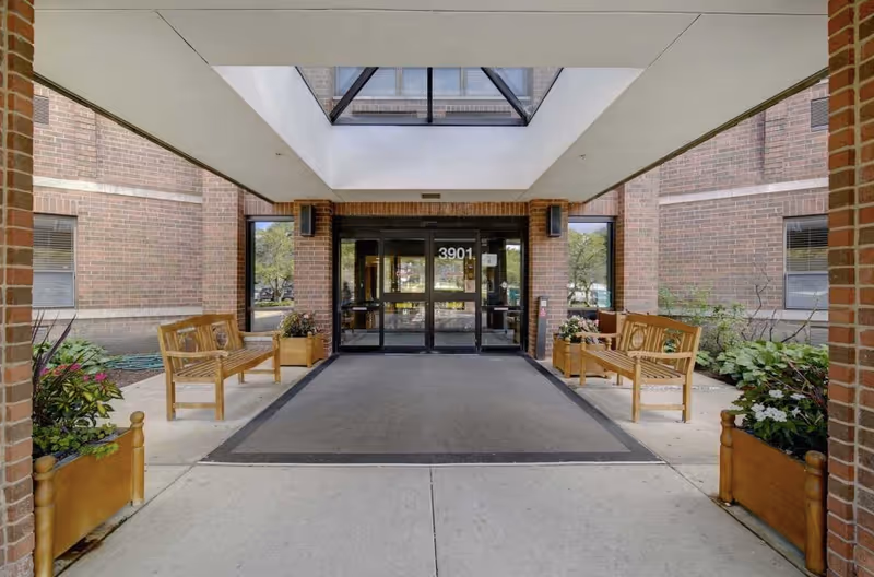 Entrance area of a brick building with glass double doors marked 3901, flanked by windows. There are wooden benches and planters with flowers on either side of the entrance under a covered walkway.