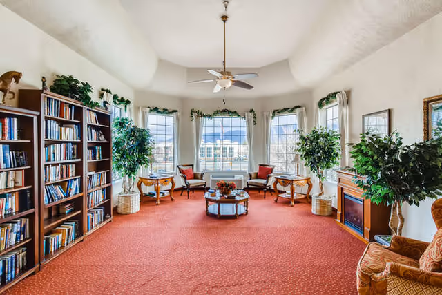 Bright communal living room with bookshelves, seating around a coffee table, large windows and indoor plants.