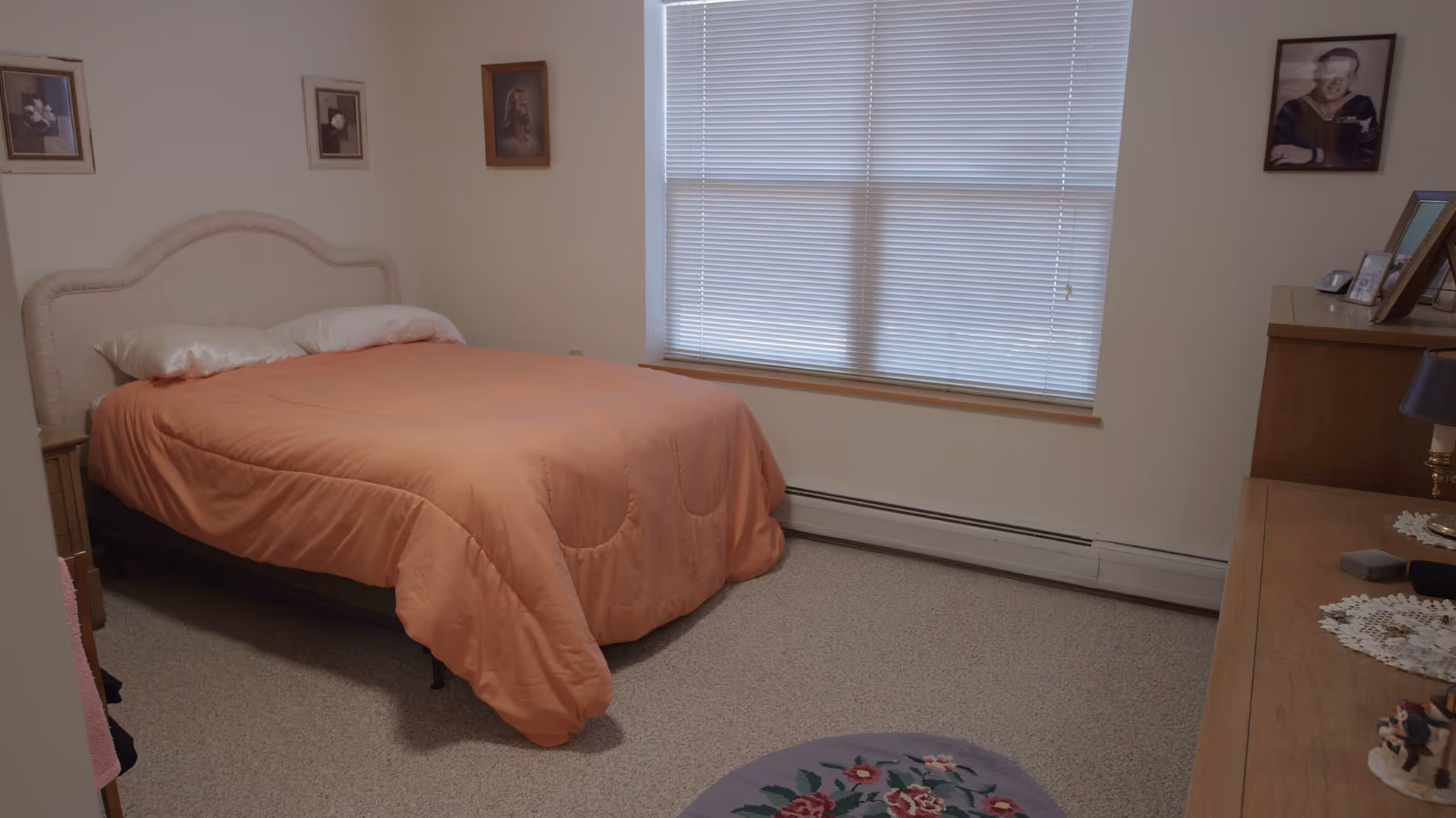 A bedroom with a bed covered in a peach-colored comforter and two white pillows. The room has a large window with closed blinds, a wooden dresser with framed photos and a lamp, and three framed pictures hanging on the walls. The floor is carpeted with a small floral rug near the dresser.