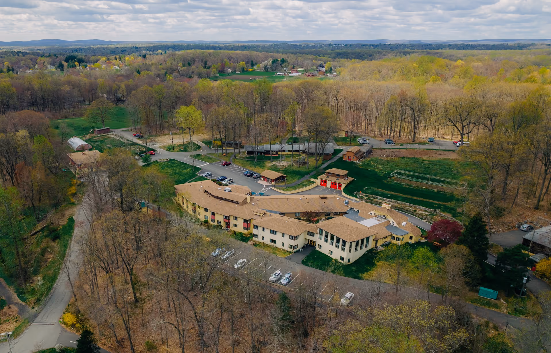 Aerial view of The Fellowship Community facility surrounded by trees and greenery. The building has a tan roof and light-colored walls, with parking lots and pathways around it. The landscape includes wooded areas and open grassy fields under a partly cloudy sky.