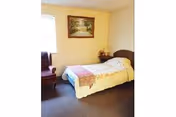 Small single-occupancy bedroom with a twin bed covered by a quilt, an armchair by the window, and a framed landscape painting on the wall.