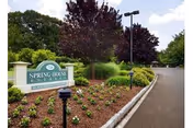 Entrance sign for Spring House Estates surrounded by landscaped flower beds and greenery along a paved road under a partly cloudy sky.