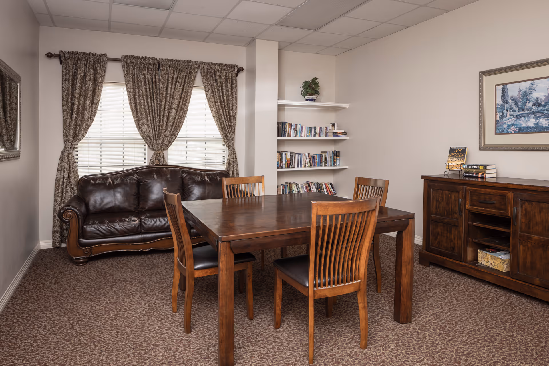 A cozy room with a dark brown leather sofa against a window with patterned curtains. In front of the sofa is a wooden dining table with four matching chairs. To the right, there is a wooden sideboard with books and a framed picture hanging above it. A small bookshelf with books and a plant is built into the corner wall.