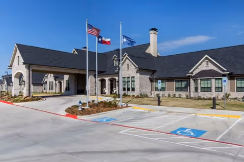 Exterior view of a single-story senior living facility building with a stone facade and dark shingled roof under a clear blue sky. Three flagpoles with the American flag, Texas state flag, and another flag stand in front of the building. There is a driveway and several handicap parking spaces in the foreground.