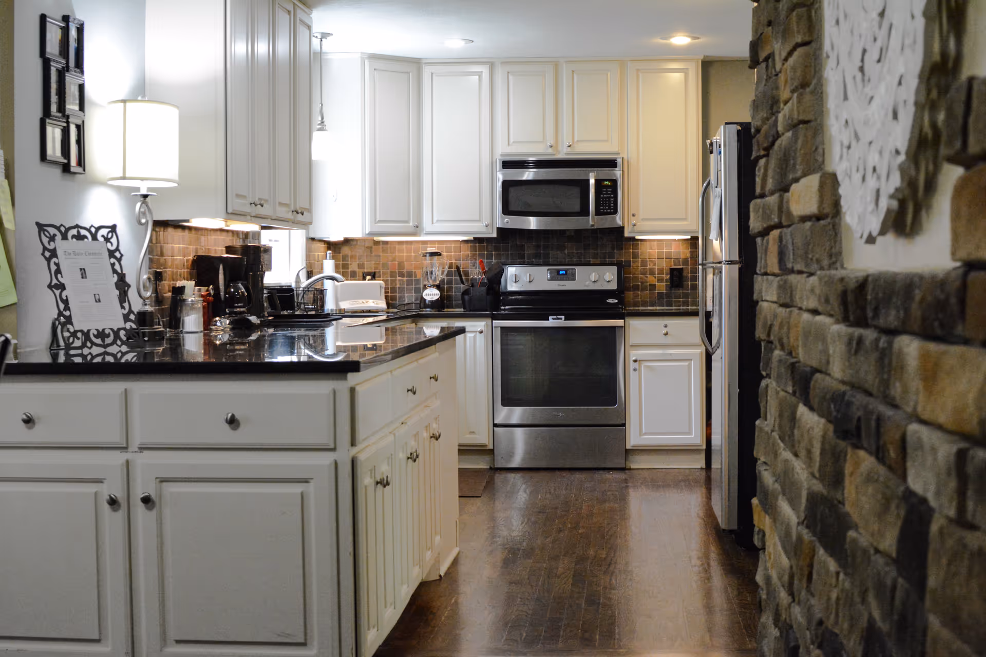 A modern kitchen with white cabinets, a black countertop, stainless steel appliances including a stove, microwave, and refrigerator. There is a stone accent wall on the right side and a lamp on the left countertop.