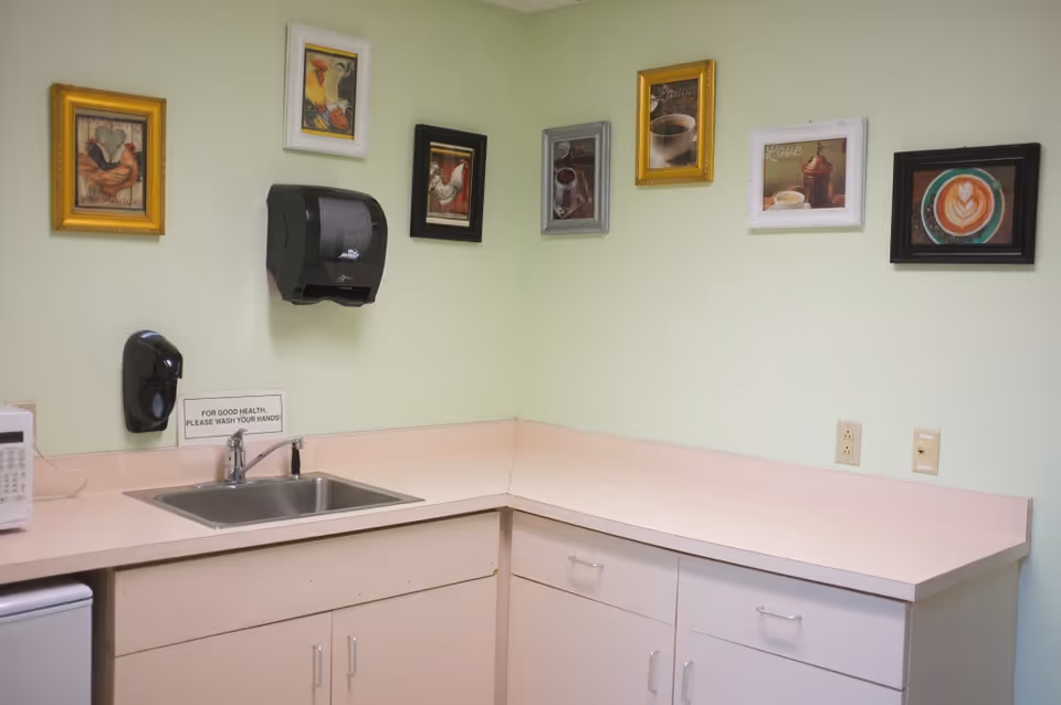 A small kitchen area with a stainless steel sink, light pink countertops, and white cabinets. Above the sink, there is a black paper towel dispenser and a soap dispenser mounted on a light green wall. Several framed pictures of roosters and coffee-themed images are hung on the walls. A microwave and a mini refrigerator are partially visible on the left side. A sign near the sink reads 'FOR GOOD HEALTH, PLEASE WASH YOUR HANDS!'