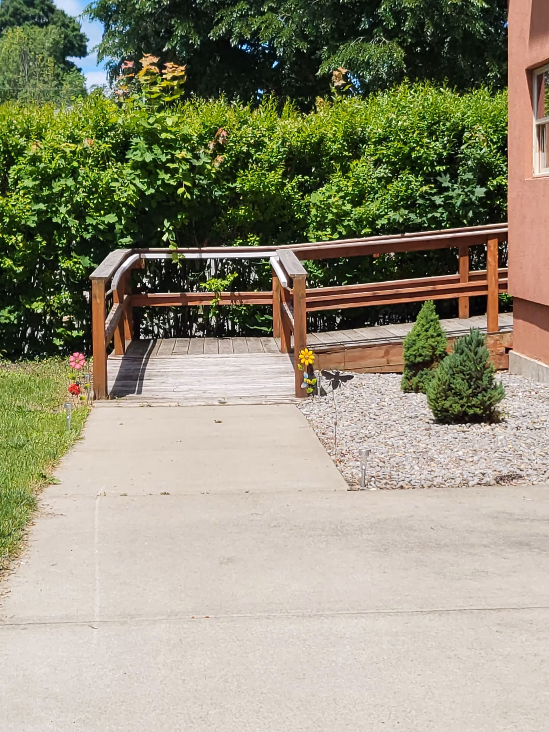 A wooden wheelchair ramp leading up to a building entrance, surrounded by green bushes and small decorative plants on a bed of rocks, with a concrete pathway in the foreground.