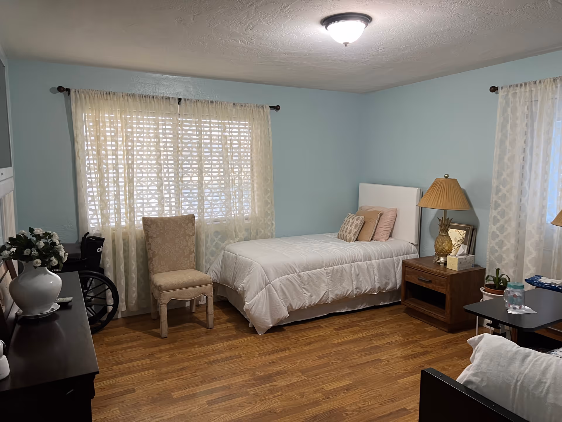 Bedroom in a senior living facility featuring a bed, chair, nightstand, lamp, and windows.