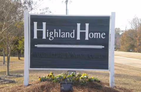 Exterior sign reads Highland Home with flowers at the base by the road.