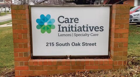 A brick-framed sign outdoors displaying the name and address of a facility: Care Initiatives Lamoni Specialty Care, 215 South Oak Street. There is a logo with overlapping blue and green shapes to the left of the text.