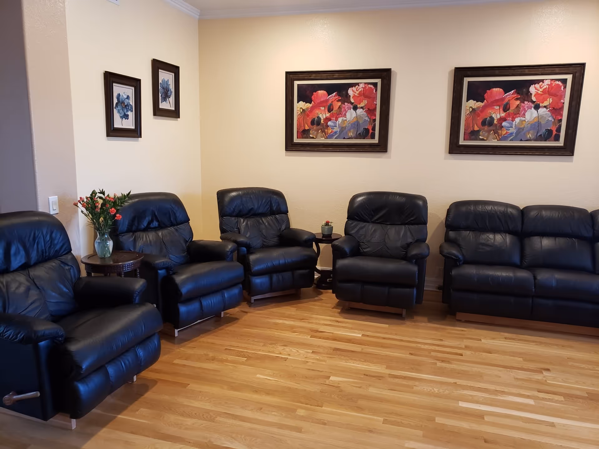A living room with five black leather recliner chairs and one black leather sofa arranged against beige walls. There are two small round wooden side tables, one with a vase of flowers and the other with a small potted plant. The walls are decorated with framed floral paintings, including two smaller blue flower paintings on the left wall and two larger colorful flower paintings on the back wall. The floor is light wood.