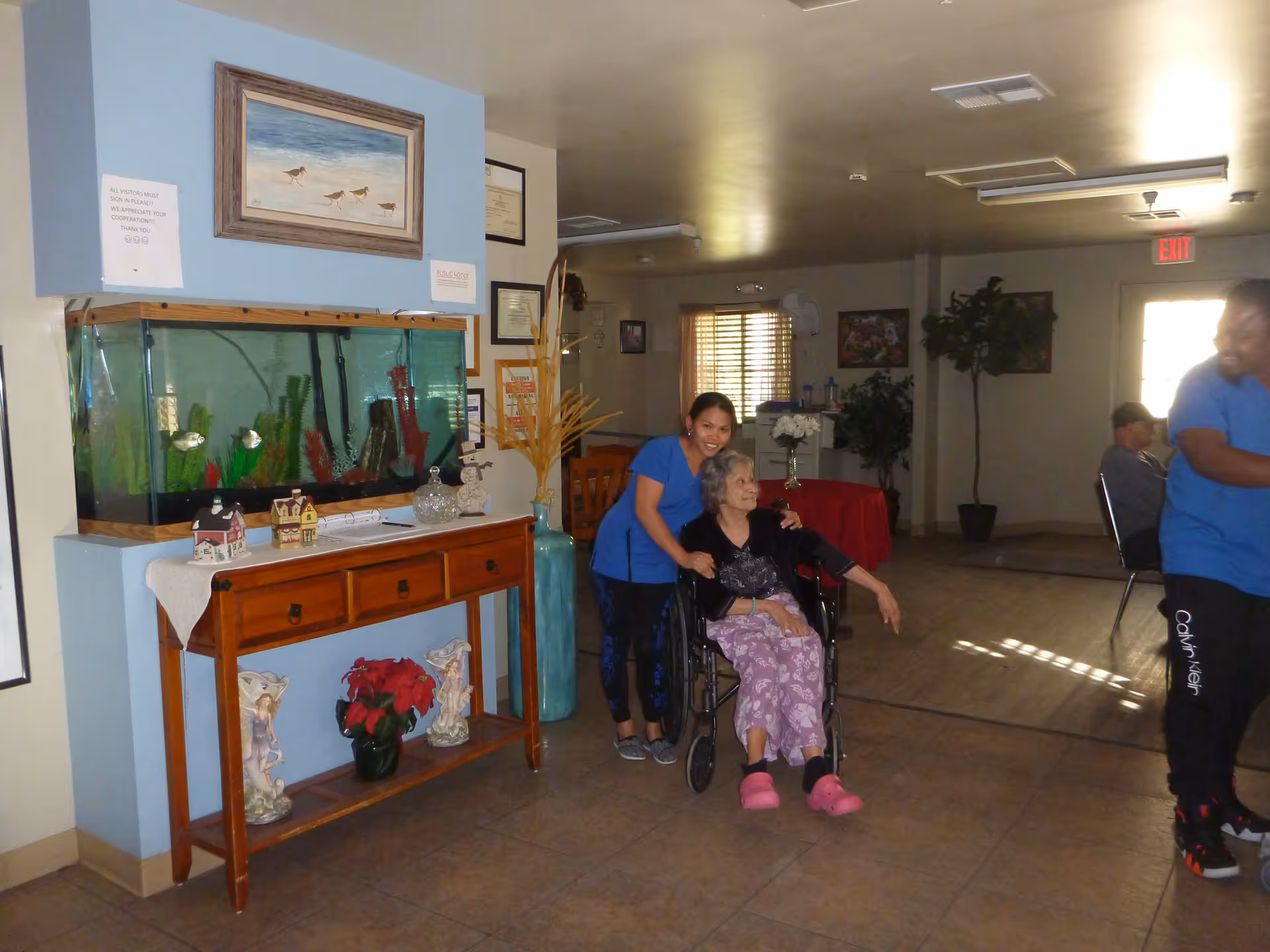 An elderly woman in a wheelchair is accompanied by a caregiver in a blue uniform inside a common area of a senior living facility. The room features a wooden table with a large fish tank on top, decorative items, and framed pictures on the wall. Another person in a blue uniform is partially visible on the right side, and a person is seated in the background near a window with blinds.