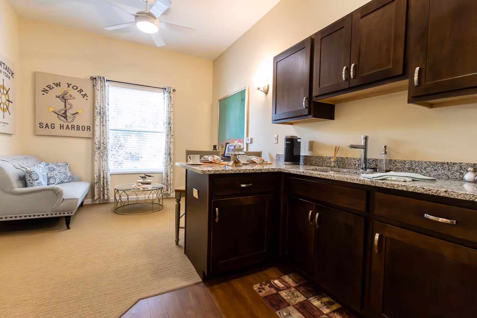 Interior view of a senior living facility showing a small kitchen area with dark wooden cabinets and granite countertops on the right, and a cozy living area with a light gray couch, decorative pillows, a round glass coffee table, and a window with patterned curtains on the left. Nautical-themed wall art is visible above the couch.
