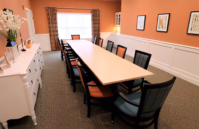 Long rectangular dining table surrounded by chairs in a warmly lit orange-walled dining/meeting room with a sideboard and framed art.