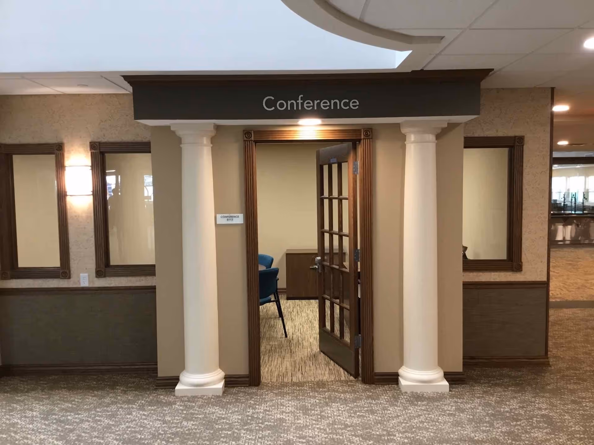 Entrance to a conference room with an open wooden door framed by two white columns. The word 'Conference' is displayed above the door. The surrounding walls have beige wallpaper and decorative wooden trim, with carpeted flooring extending into the room.