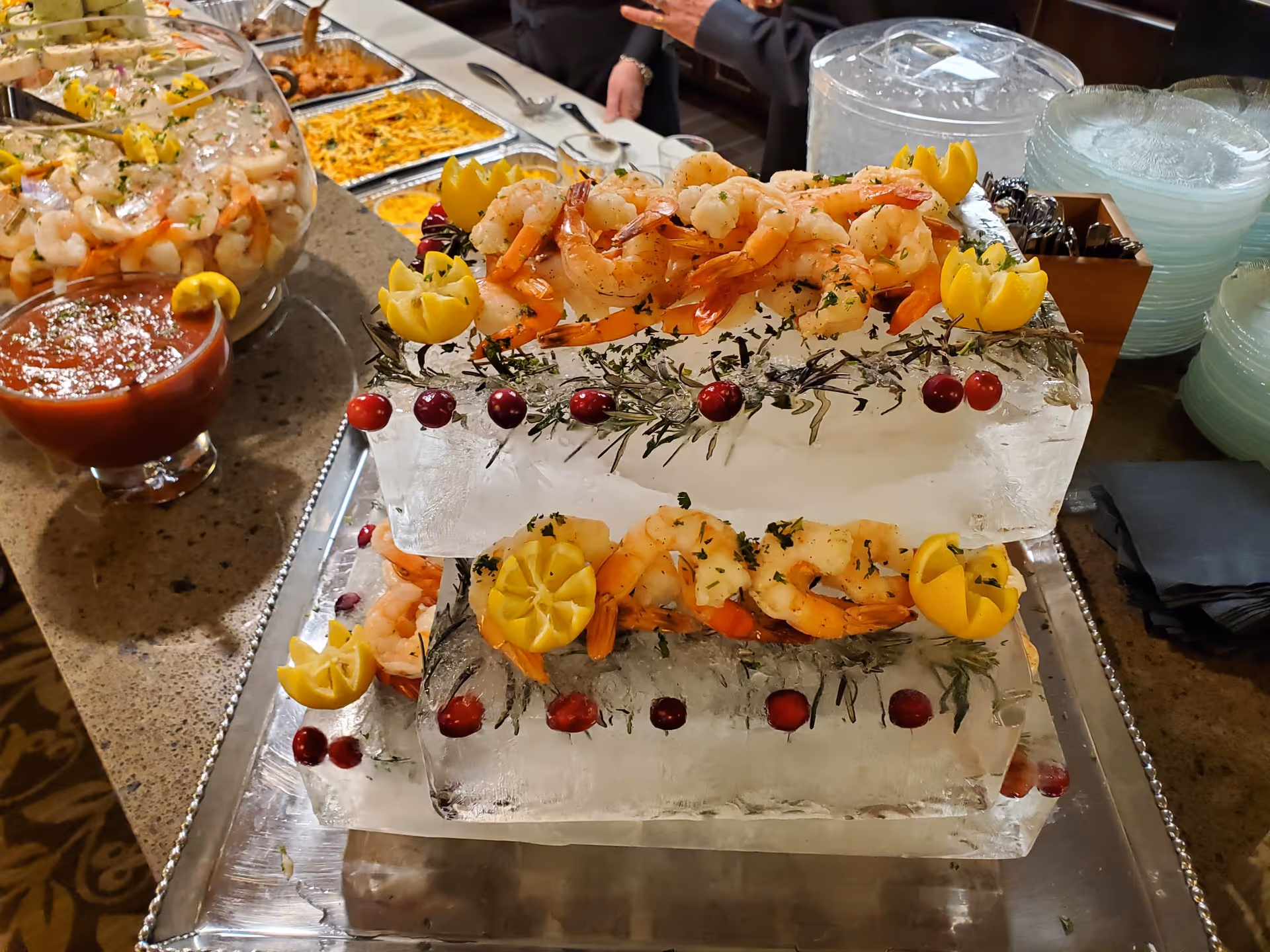 A seafood buffet display featuring shrimp arranged on two large blocks of ice decorated with lemon wedges, herbs, and cranberries. A bowl of cocktail sauce is placed nearby, along with plates and utensils in the background.