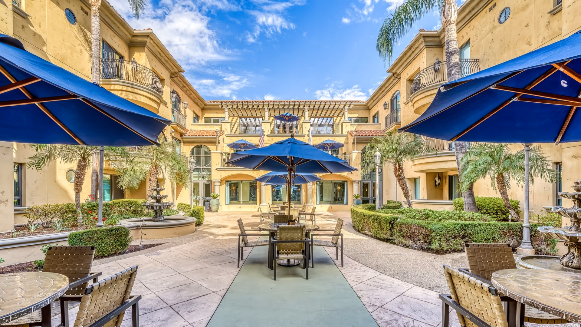 Outdoor courtyard area at Meridian at Chino featuring multiple tables with blue umbrellas, surrounded by palm trees, shrubs, and a beige multi-story building under a partly cloudy sky.