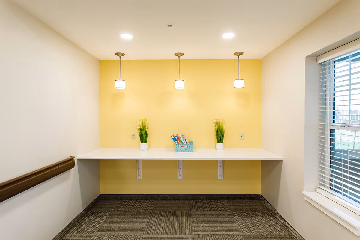 A small, brightly lit nook with a white countertop mounted against a yellow accent wall. Three pendant lights hang from the ceiling above the countertop. On the countertop, there are two small potted plants with green grass-like foliage and a small blue basket containing some items. To the right, there is a window with white blinds partially open, and to the left, a wooden handrail is mounted on the wall. The floor is carpeted with a patterned design.