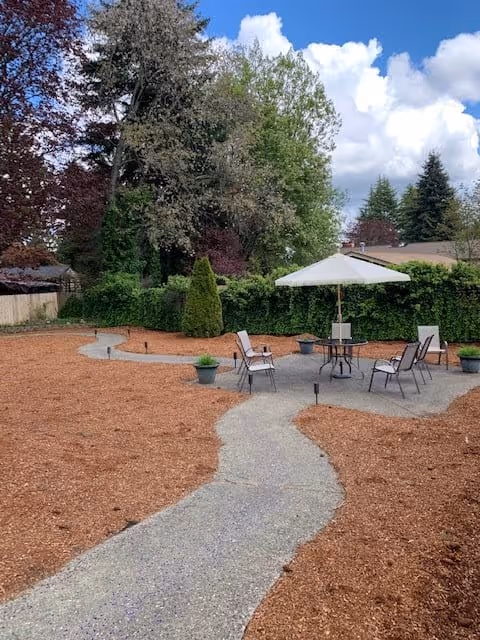 Outdoor courtyard with a winding paved path leading to a patio seating area with a table, chairs, and umbrella surrounded by mulch and trees.