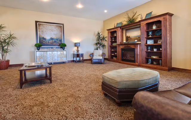 A spacious living room with patterned carpet, a large wooden entertainment center with a fireplace, shelves decorated with various items, a framed painting above a console table, two armchairs, a coffee table, and potted plants.