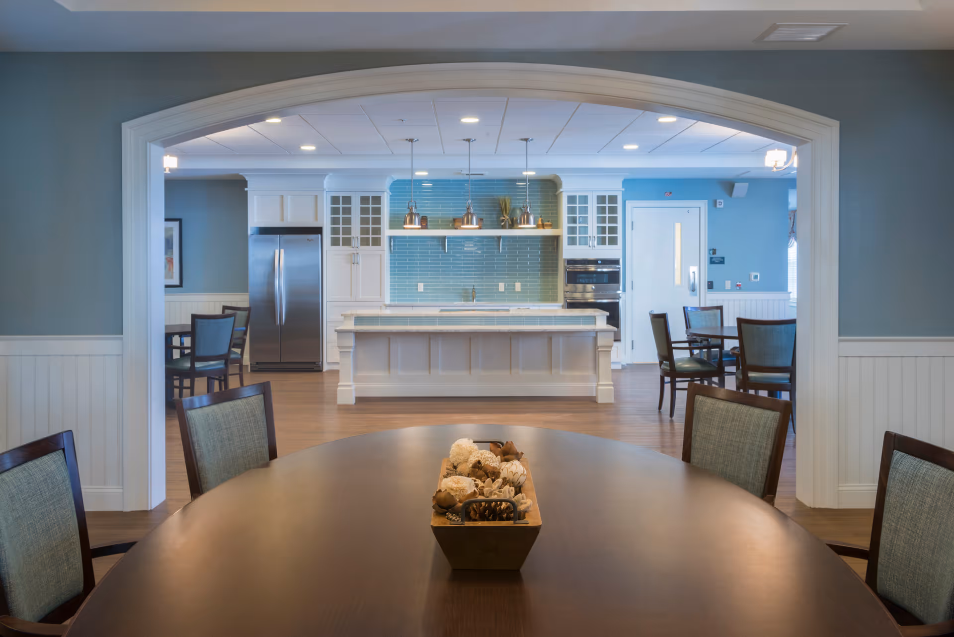 View of a senior living facility's dining area with a round wooden table and chairs in the foreground, leading into a kitchen area with a large island, white cabinetry, stainless steel refrigerator, and blue tiled backsplash. Pendant lights hang above the island, and additional dining tables and chairs are visible in the background.