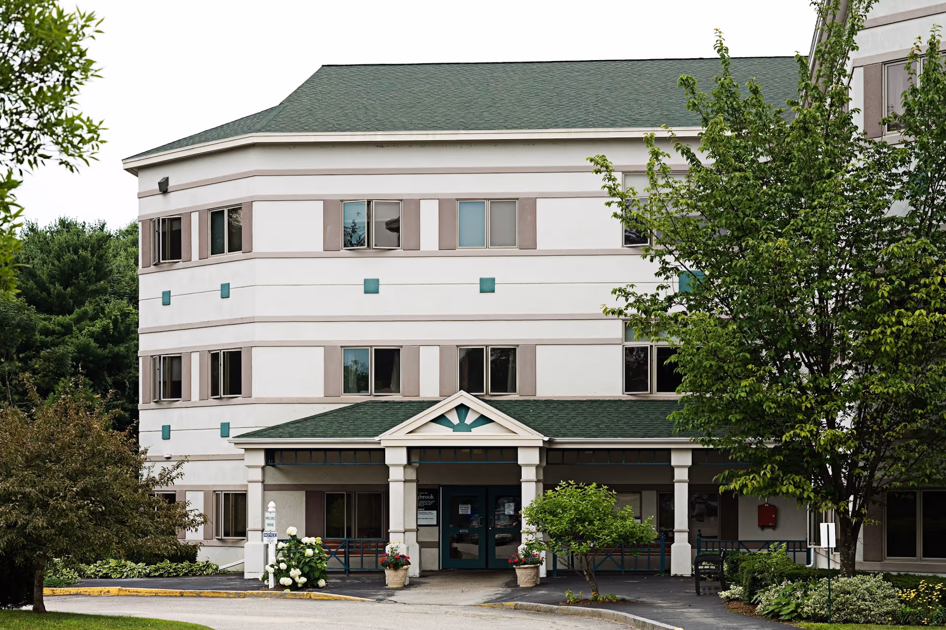 Front exterior of a multi-story senior living building with a covered entrance, planters, and trees.