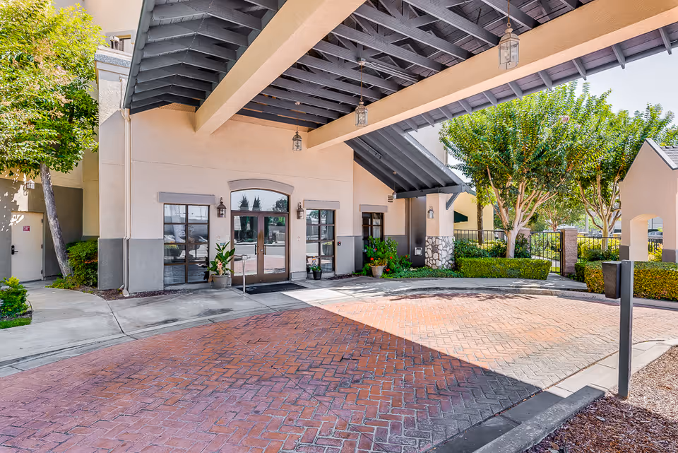 Entrance area of Westminster Terrace Senior Living with a covered driveway featuring a brick-patterned pavement, beige walls with gray trim, glass double doors, potted plants, and trees with green foliage surrounding the entrance.