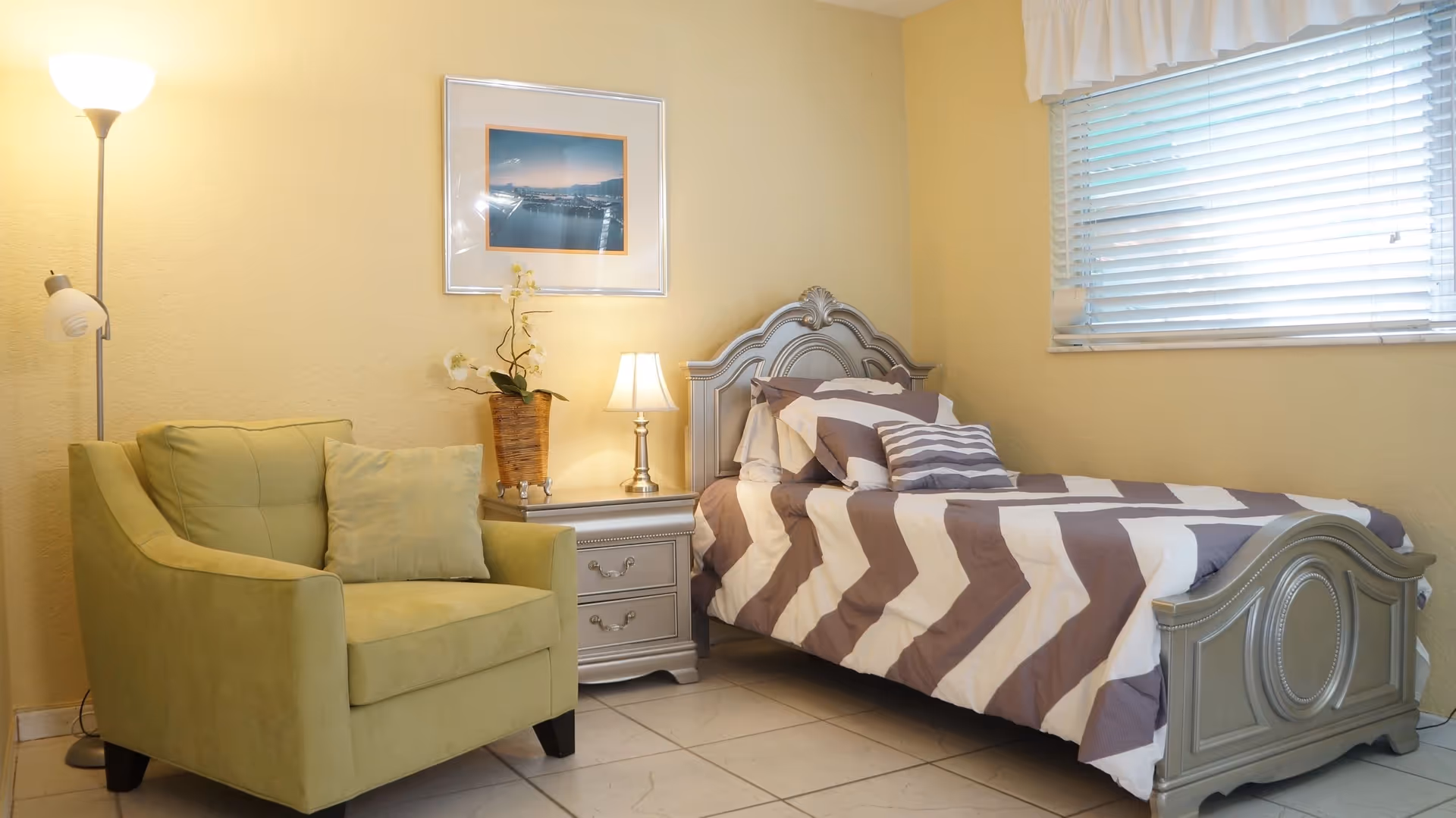 A cozy bedroom with a single bed featuring a gray and white chevron patterned comforter, a matching headboard and footboard, a nightstand with a small lamp and a potted orchid, a green armchair with a matching pillow, a floor lamp, and a window with white blinds and a valance.