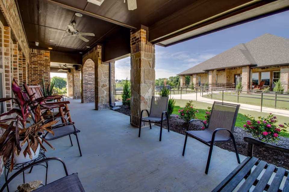 Covered outdoor patio with chairs and ceiling fans overlooking a landscaped courtyard and neighboring building.