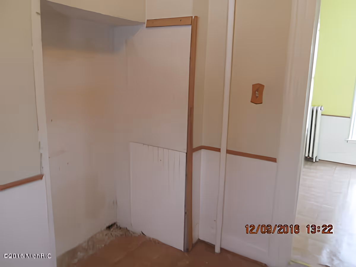 An empty corner of a room with white walls and brown trim. The lower part of the walls has paneling, and there is some visible damage or wear near the floor. A doorway leads to another room with a green wall and a radiator visible. The floor is bare and appears to be under renovation or in poor condition.